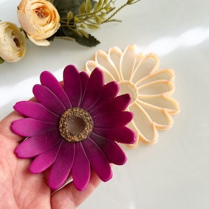 May include: A magenta daisy flower with a textured brown centre is held in a hand. A second, cream-coloured daisy is next to it. Artificial flowers and greenery are in the background. The flowers are on a white surface.