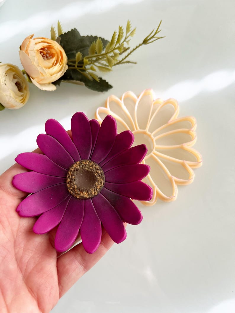 May include: A magenta daisy flower with a textured brown centre is held in a hand. A second, cream-coloured daisy is next to it. Artificial flowers and greenery are in the background. The flowers are on a white surface.