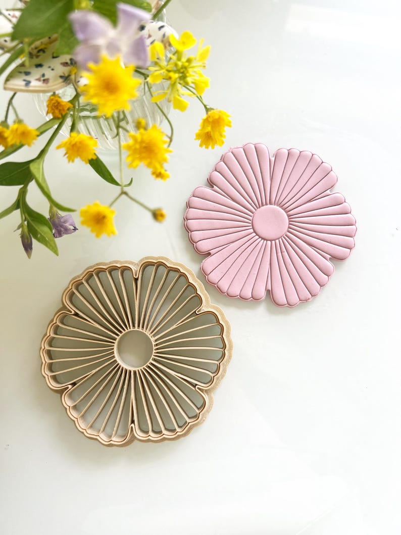 May include: Two flower-shaped items. One is a light pink, solid, and the other is a light brown, openwork design. Both have a circular center and radiating lines. Yellow flowers and greenery are in a vase in the background.