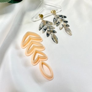 Könnte beinhalten: Ein Paar Hängeohrringe mit einem Blattdesign. Die Ohrringe sind aus weiß-schwarzem marmoriertem Harz gefertigt. Die Ohrringe hängen an einem goldfarbenen Haken. Ein Satz blattförmiger Ausstecher ist ebenfalls im Bild zu sehen.