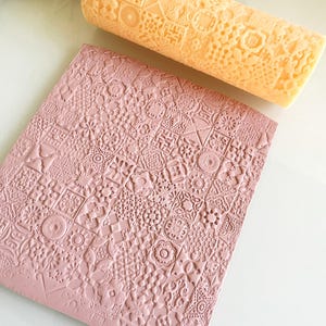 Pu&ograve; includere: Un mattarello color pesca chiaro con un motivo geometrico dettagliato &egrave; appoggiato sopra un foglio a motivi rosa polveroso. Il mattarello &egrave; cilindrico e il foglio &egrave; rettangolare. I motivi includono quadrati, cerchi e motivi floreali.