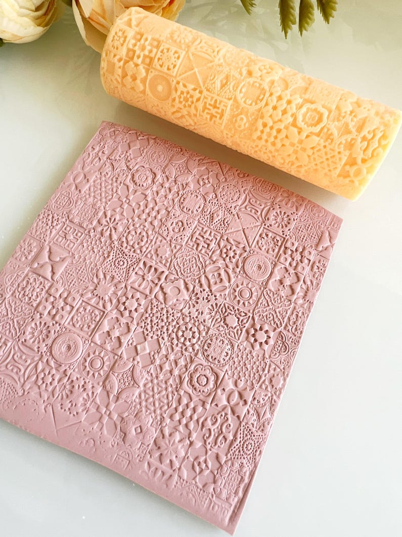 Pu&ograve; includere: Un mattarello color pesca chiaro con un motivo geometrico dettagliato &egrave; appoggiato sopra un foglio a motivi rosa polveroso. Il mattarello &egrave; cilindrico e il foglio &egrave; rettangolare. I motivi includono quadrati, cerchi e motivi floreali.