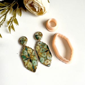 Puede incluir: Pendientes artesanales con diseño floral en tonos verdes, azules y amarillos, con detalles en metal dorado. La imagen también muestra un anillo y un molde, todo sobre un fondo blanco.