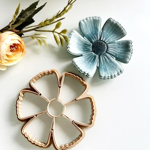 May include: A light blue flower-shaped cookie cutter and a blue flower-shaped confection. The cookie cutter is made of a light brown material. A peach-coloured flower and green leaves are in the background. The items are on a white surface.