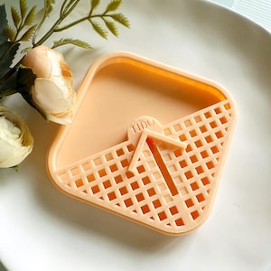 May include: A peach-colored, square soap dish with a lattice design on one half and the word "TIDO" printed on the top. The dish sits on a white plate, with a sprig of greenery and a cream-colored rose in the background.
