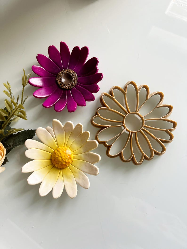 May include: Three daisy-shaped items. One is a vibrant magenta with a brown centre, another is a light tan outline, and the third is a white and yellow daisy with a textured yellow centre. Floral theme.