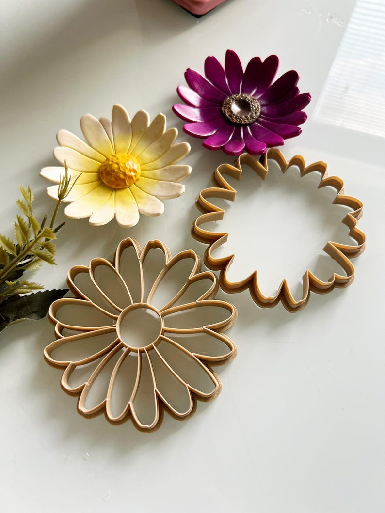 May include: Four daisy-shaped items are shown from above. Two are colourful, one with yellow and white petals and another with purple petals. The other two are tan-coloured daisy-shaped cookie cutters. They are arranged on a white surface.