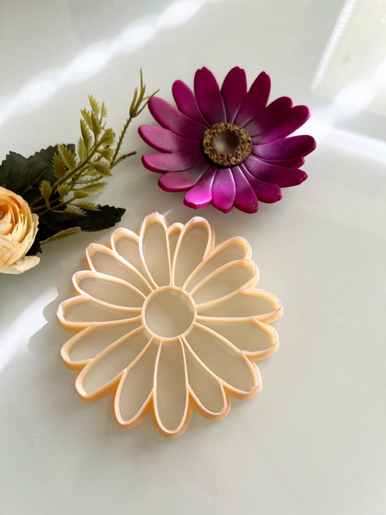May include: Two daisy-shaped objects. One is a light peach colour, with a hollow centre and petal outlines. The other is a deep magenta with a textured centre. A sprig of greenery and a peach-coloured flower are also present.