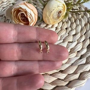 以下が含まれることがあります： 小さなゴールドカラーのフープピアス2つ。手で持たれており、背景には織りマットと造花があります。ピアスには、チャームなどを取り付けるための小さなループが下部にあります。