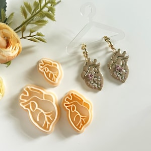 以下が含まれることがあります： ハート型のクッキーカッター3個セットと、ピンクと白の花柄の金色のハート型のイヤリングのペア。イヤリングは透明なプラスチック製のハンガーにかけられています。