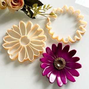 May include: A top-down view shows three flower-shaped cookie cutters in beige and a purple flower. The cookie cutters are daisy and sunflower designs. A faux floral arrangement with beige roses and green leaves is in the background.