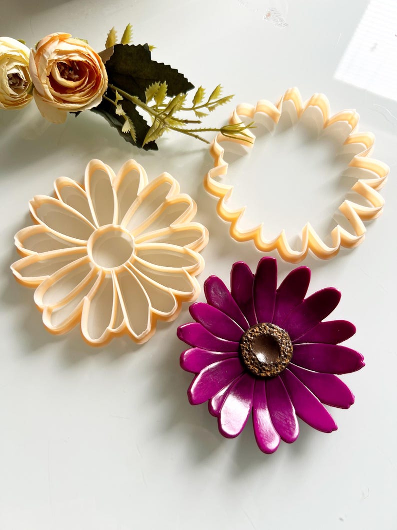 May include: A top-down view shows three flower-shaped cookie cutters in beige and a purple flower. The cookie cutters are daisy and sunflower designs. A faux floral arrangement with beige roses and green leaves is in the background.