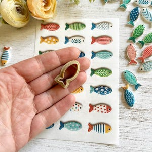 May include: A beige plastic fish-shaped cookie cutter held in front of a white sheet featuring a grid of various small, colorful, patterned fish illustrations, with several loose ceramic fish charms scattered to the right side.
