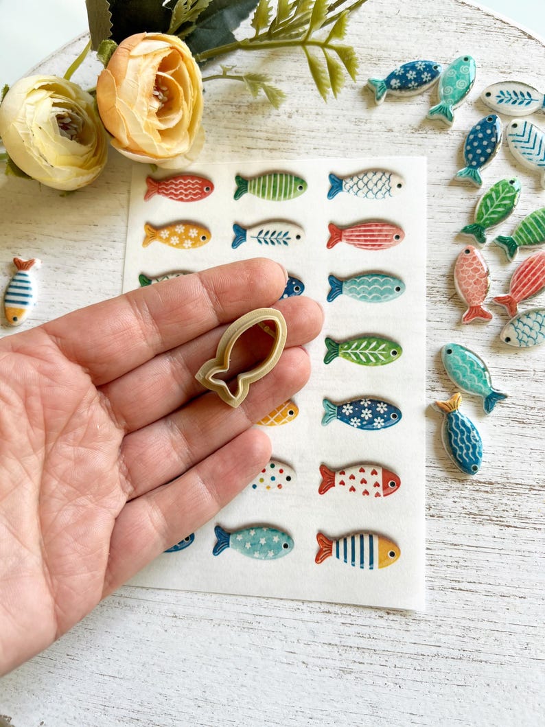 May include: A beige plastic fish-shaped cookie cutter held in front of a white sheet featuring a grid of various small, colorful, patterned fish illustrations, with several loose ceramic fish charms scattered to the right side.