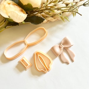 May include: A set of four plastic cookie cutters in a light brown color. The cutters are shaped like a bow, a pair of bunny ears, a single oval, and a small rectangle.