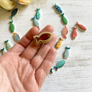 May include: A beige plastic fish-shaped cookie cutter held in a palm, surrounded by numerous small, flat, ceramic fish-shaped ornaments in various colors including blue, green, orange, and pink, each featuring distinct patterns like stripes and dots.