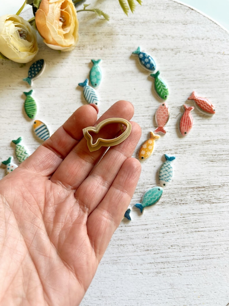 May include: A beige plastic fish-shaped cookie cutter held in a palm, surrounded by numerous small, flat, ceramic fish-shaped ornaments in various colors including blue, green, orange, and pink, each featuring distinct patterns like stripes and dots.