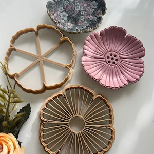 May include: Four flower-shaped items are displayed. One is a floral-patterned dish, another is a pink dish with a central design, and two are wooden cookie cutters. The items are arranged on a white surface, with a few flowers in the lower left corner.