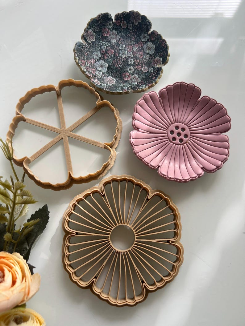 May include: Four flower-shaped items are displayed. One is a floral-patterned dish, another is a pink dish with a central design, and two are wooden cookie cutters. The items are arranged on a white surface, with a few flowers in the lower left corner.
