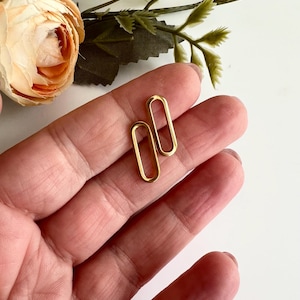 以下が含まれることがあります： 2つの金色の、開いた楕円形のイヤリング。イヤリングは人の手で持たれており、背景には花と緑がぼやけています。イヤリングはシンプルでエレガントで、普段使いに適しています。