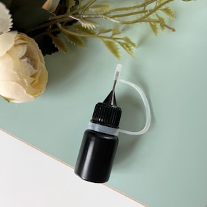 Puede incluir: Una pequeña botella negra con una tapa negra y un aplicador de plástico blanco. La botella es cilíndrica y parece contener un líquido. El fondo es de color verde claro con algunos acentos florales.