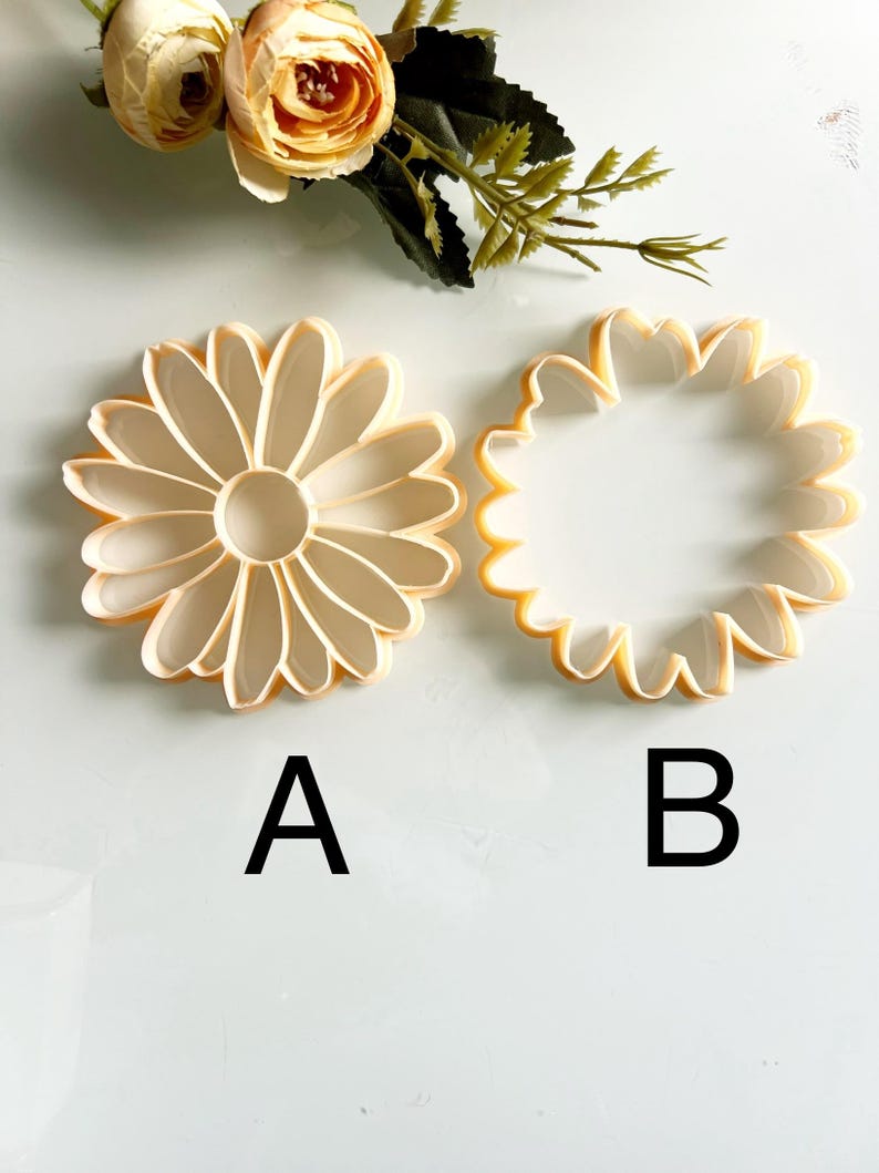 May include: Two daisy-shaped cookie cutters, labelled A and B, in light yellow and white. The cutters are made of plastic and are designed to cut out flower shapes. Artificial flowers are in the background.