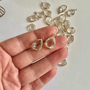 Puede incluir: Primer plano de una mano sosteniendo dos colgantes de cristal transparente en forma de lágrima con bucles de metal dorado. Los colgantes están rodeados de una dispersión de colgantes similares sobre una superficie blanca.