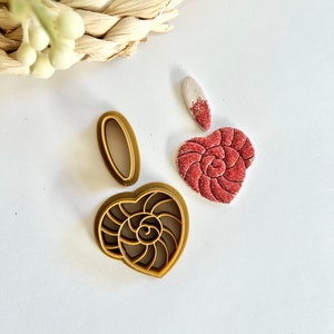 May include: A set of two brown plastic cutters featuring an oval shape and a heart-shaped nautilus shell design, alongside a finished red textured heart-shaped clay piece with a small white and red speckled teardrop-shaped clay attachment.