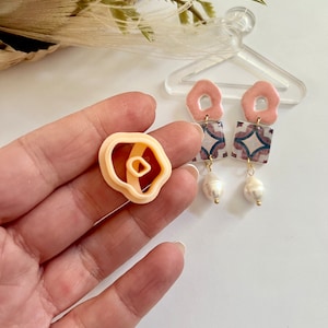 Puede incluir: Un par de pendientes de color rosa y blanco con un patrón geométrico y detalles de perlas. Los pendientes se muestran sobre una superficie blanca con un colgador de plástico transparente.