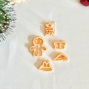 Puede incluir: Un conjunto de cortadores de galletas de plástico beige con formas variadas, como un hombre de jengibre, un vestido, un sombrero y una camisa. Los cortadores están dispuestos sobre una superficie blanca, ideales para hornear en Navidad. También se incluye un cortador rectangular con un diseño.