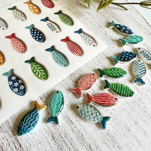 May include: A collection of small, flat, fish-shaped ceramic ornaments featuring various patterns and colors including blue, green, red, and orange. Some fish are arranged on a white sheet, while others are scattered, each with a small hole.