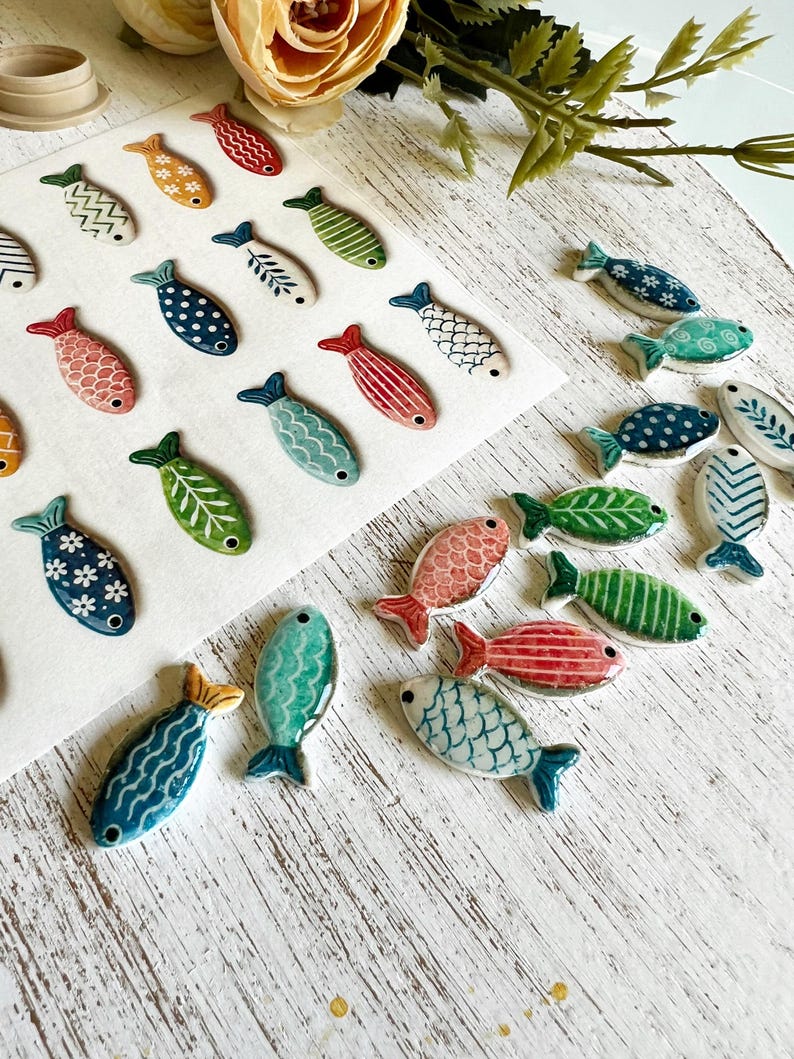 May include: A collection of small, flat, fish-shaped ceramic ornaments featuring various patterns and colors including blue, green, red, and orange. Some fish are arranged on a white sheet, while others are scattered, each with a small hole.