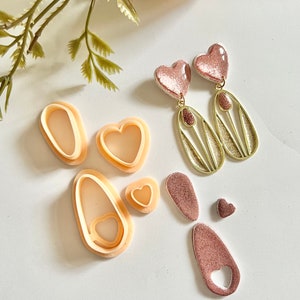 以下が含まれることがあります： ハート型と楕円形のポリマークレイイヤリングモールドのセット。モールドはベージュで、滑らかな仕上がりです。画像には、完成したイヤリングが2組あり、1組はゴールドのアクセントがついたピンク、もう1組はゴールドのアクセントがついた薄い茶色です。