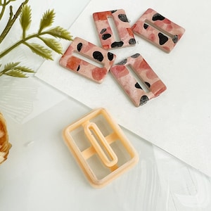 May include: A collection of handmade polymer clay rectangle-shaped earrings with a cutout design. The earrings feature a speckled pattern in shades of pink, red, and black. A light orange earring cutter is also present.