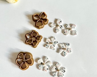 Peut inclure: Un ensemble de six boucles d'oreilles en forme de fleur. Les boucles d'oreilles sont en céramique blanche avec un motif moucheté et des accents dorés. Les boucles d'oreilles sont assorties à des emporte-pièces en forme de fleur en plastique brun.