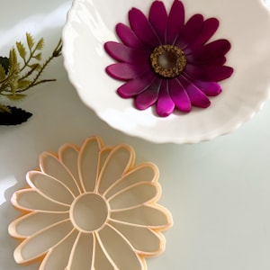 May include: A white, fluted bowl contains a bright magenta flower with a gold centre. A daisy-shaped cookie cutter in a pale peach colour is below the bowl. The objects are on a light-coloured surface.