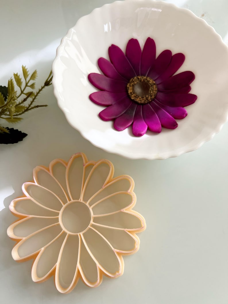 May include: A white, fluted bowl contains a bright magenta flower with a gold centre. A daisy-shaped cookie cutter in a pale peach colour is below the bowl. The objects are on a light-coloured surface.