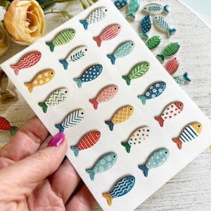 May include: A collection of small, flat, fish-shaped ceramic cabochons arranged on a white rectangular card. Each fish features a unique hand-painted pattern, including stripes, polka dots, scales, and floral motifs in various colors like blue, red, green, and yellow.