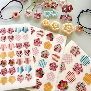 Peut inclure: Une collection d'accessoires en forme de fleur comprenant des élastiques à cheveux, des bracelets de perles et des breloques, ainsi que trois feuilles d'autocollants floraux avec des motifs variés comme des rayures, des carreaux et des fleurs.