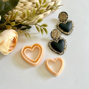 Op de afbeelding: Zwarte hartvormige oorbellen met gouden glitterrand en een bijpassende gouden glittertop. Er zijn ook twee hartvormige koekjessnijders aanwezig. De items zijn gerangschikt op een wit oppervlak met een geweven mand en bloemaccenten.