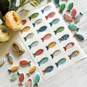May include: A collection of small, flat, fish-shaped ceramic or clay pieces in various colors and patterns, including stripes, dots, and floral motifs. Some pieces are arranged on a white rectangular sheet, while others are scattered nearby.