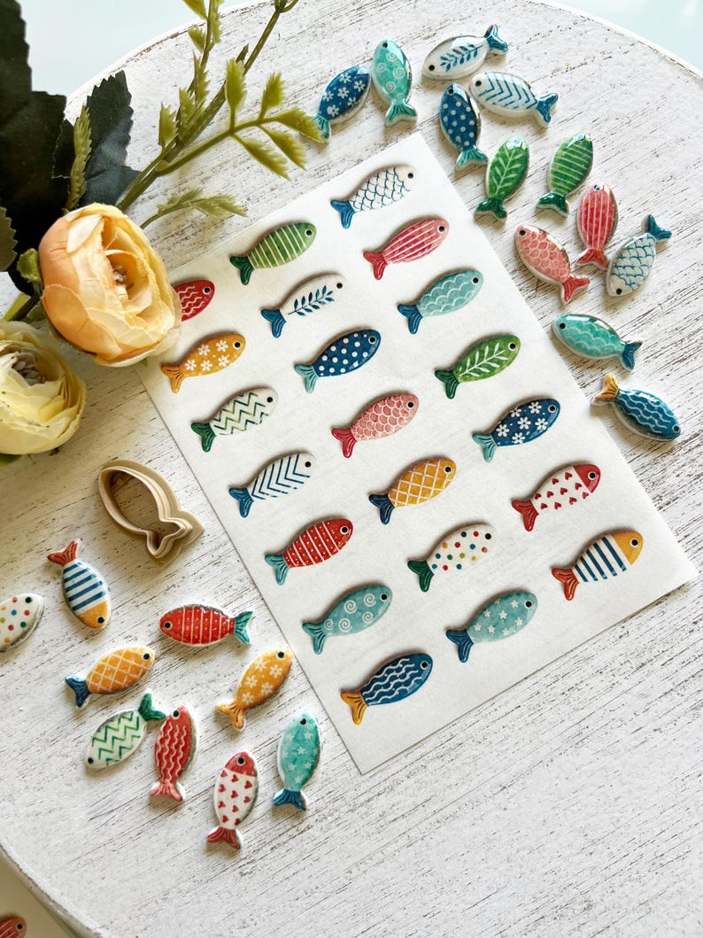 May include: A collection of small, flat, fish-shaped ceramic or clay pieces in various colors and patterns, including stripes, dots, and floral motifs. Some pieces are arranged on a white rectangular sheet, while others are scattered nearby.
