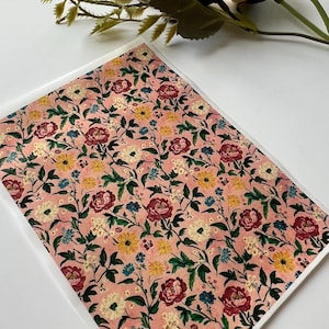 May include: A sheet of paper with a floral pattern. The design features a repeating pattern of red, yellow, white, and blue flowers with green leaves on a pink background. The paper is rectangular and appears to be a decorative element.