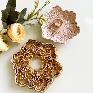 May include: A light pink, floral-patterned ring dish holds two gold rings. A matching brown floral-shaped object sits below. Artificial flowers and greenery are in the background. The dish and object have a detailed, embossed design.