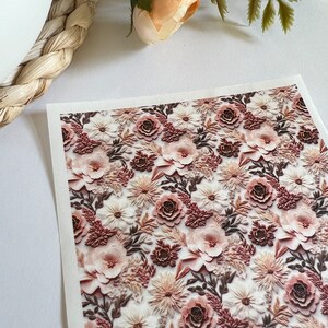 Könnte beinhalten: Ein Blatt Dekorpapier mit einem sich wiederholenden Muster aus rosa, weißen und braunen 3D-Blumen. Die Blumen sind in einem zufälligen Muster angeordnet und haben eine strukturierte Oberfläche.