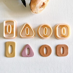 Puede incluir: Conjunto de seis cortadores de galletas en forma de rectángulo, triángulo y círculo. Cada cortador presenta una letra "O" recortada en el centro. Los cortadores se muestran junto a seis galletas cortadas con las mismas formas y letras.