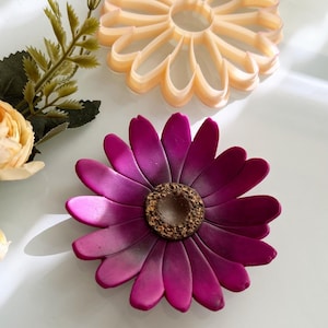 May include: Two flower-shaped objects are displayed. The lower flower is a deep magenta with a textured brown centre. The upper flower is a light peach colour and has a cutout design. The objects are on a white surface.