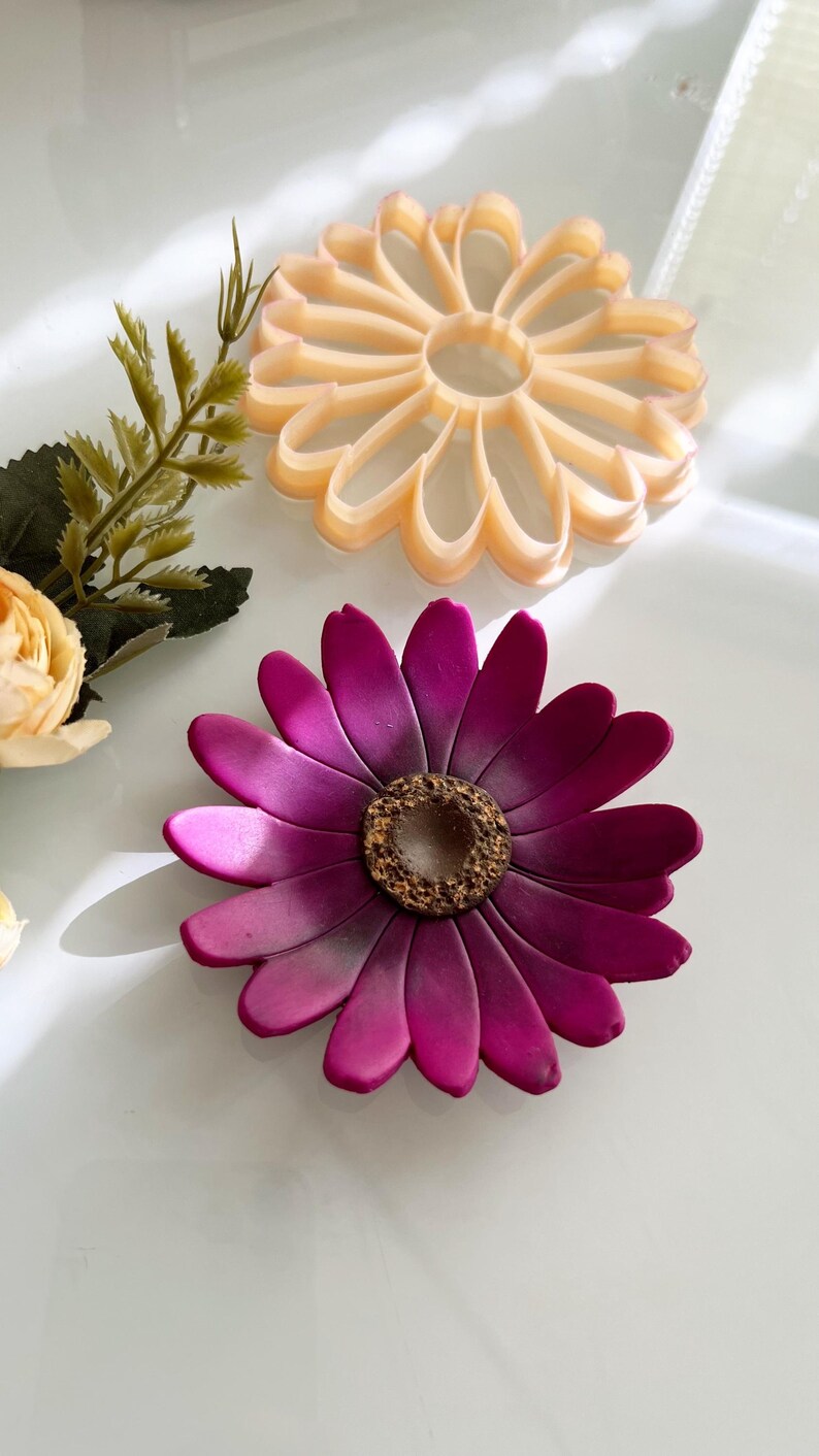 May include: Two flower-shaped objects are displayed. The lower flower is a deep magenta with a textured brown centre. The upper flower is a light peach colour and has a cutout design. The objects are on a white surface.
