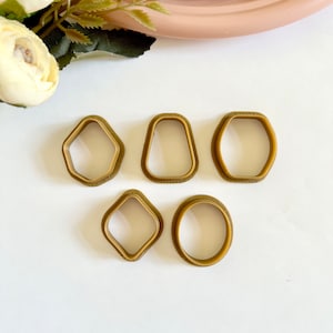 Op de afbeelding: Set van zes goudkleurige koekjesvormen in verschillende vormen, waaronder een cirkel, een ovaal, een ruit en twee verschillende zeshoeken.