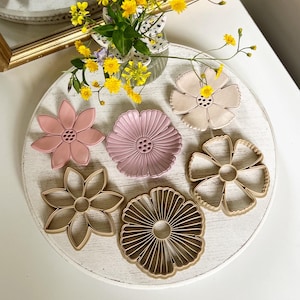 May include: A collection of flower-shaped items in various colors, including pink and beige, arranged on a white, round surface. The items are displayed with a vase of yellow flowers and a mirror in the background. The items appear to be decorative or for crafting.